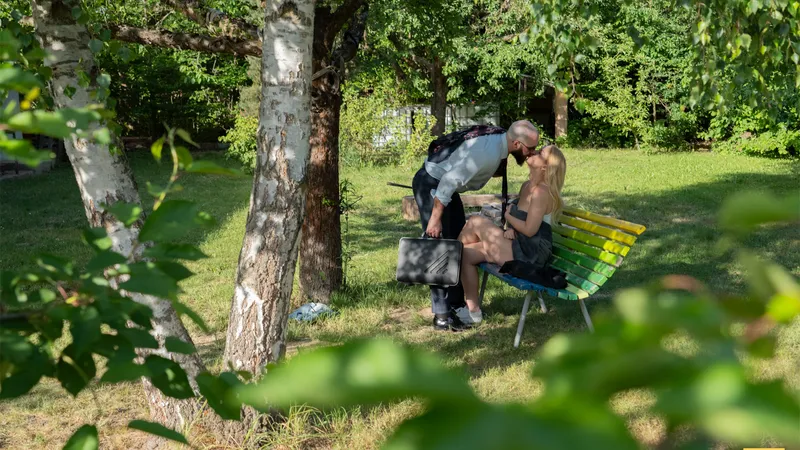 The husband leans down to kiss Millie on the bench, unaware of who is watching from the trees